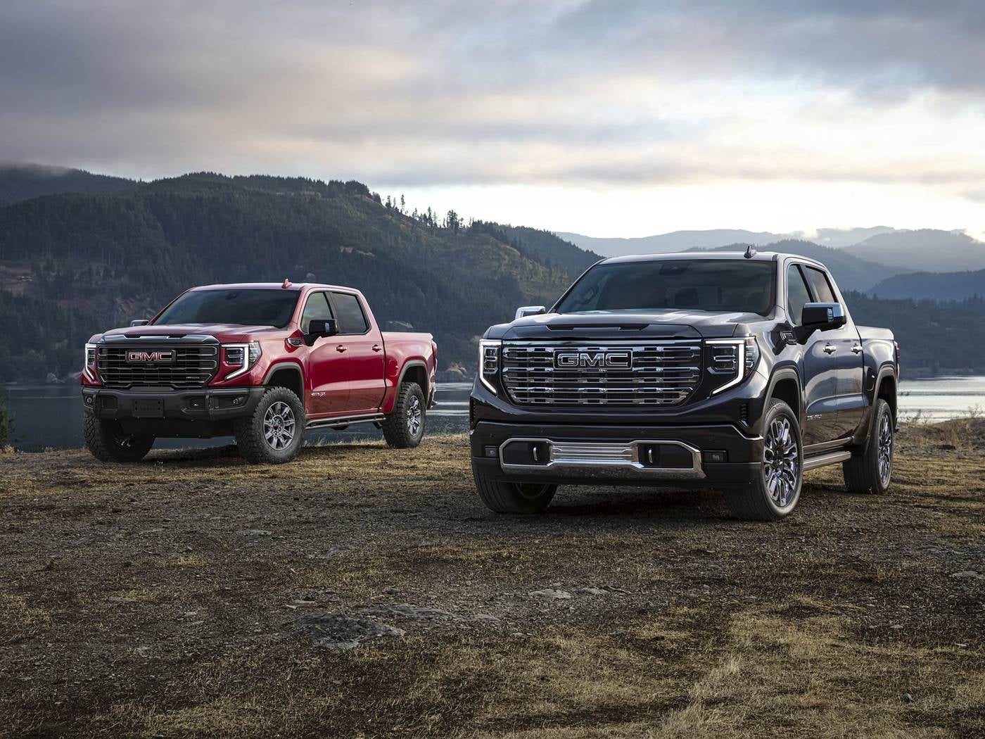 One red and one black GMC Sierra 1500 pickup parked in a grassy field beside the water